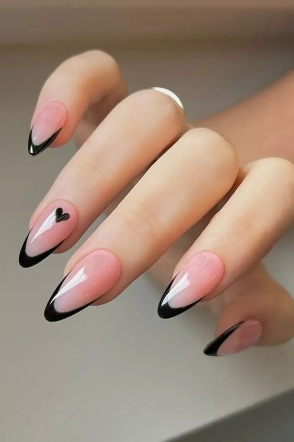 Close-up of almond-shaped nails with a sheer pink base and sharp black French tips, featuring a tiny black heart accent on one finger.
