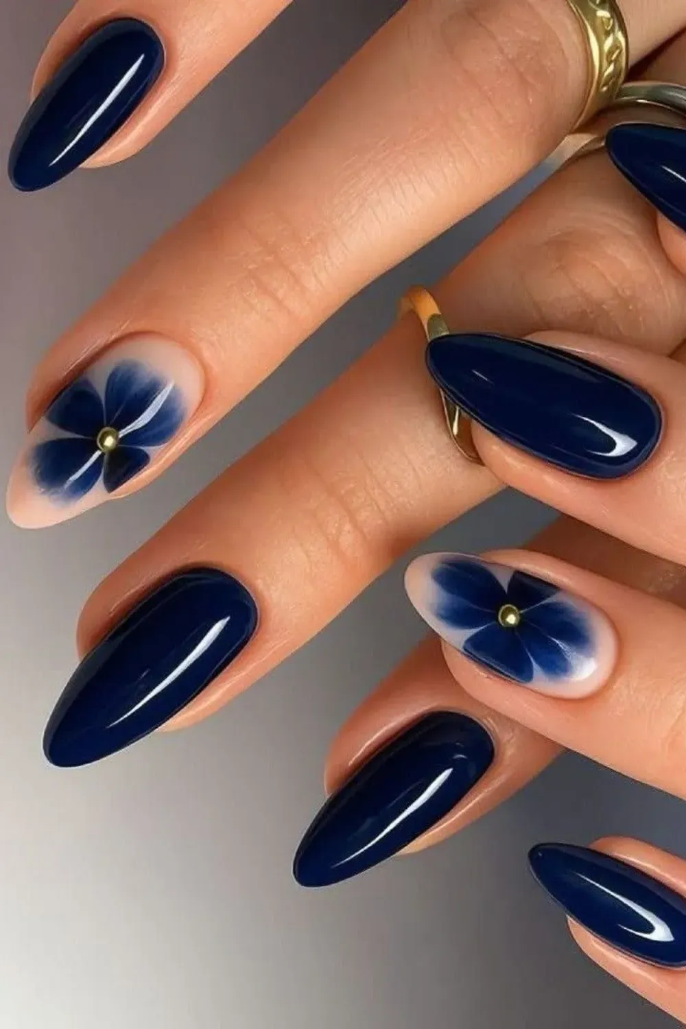 Glossy Navy Nails with Blue Floral Accent Hands with glossy, dark navy blue almond nails, featuring a single accent nail with a large blue floral design on a nude base.