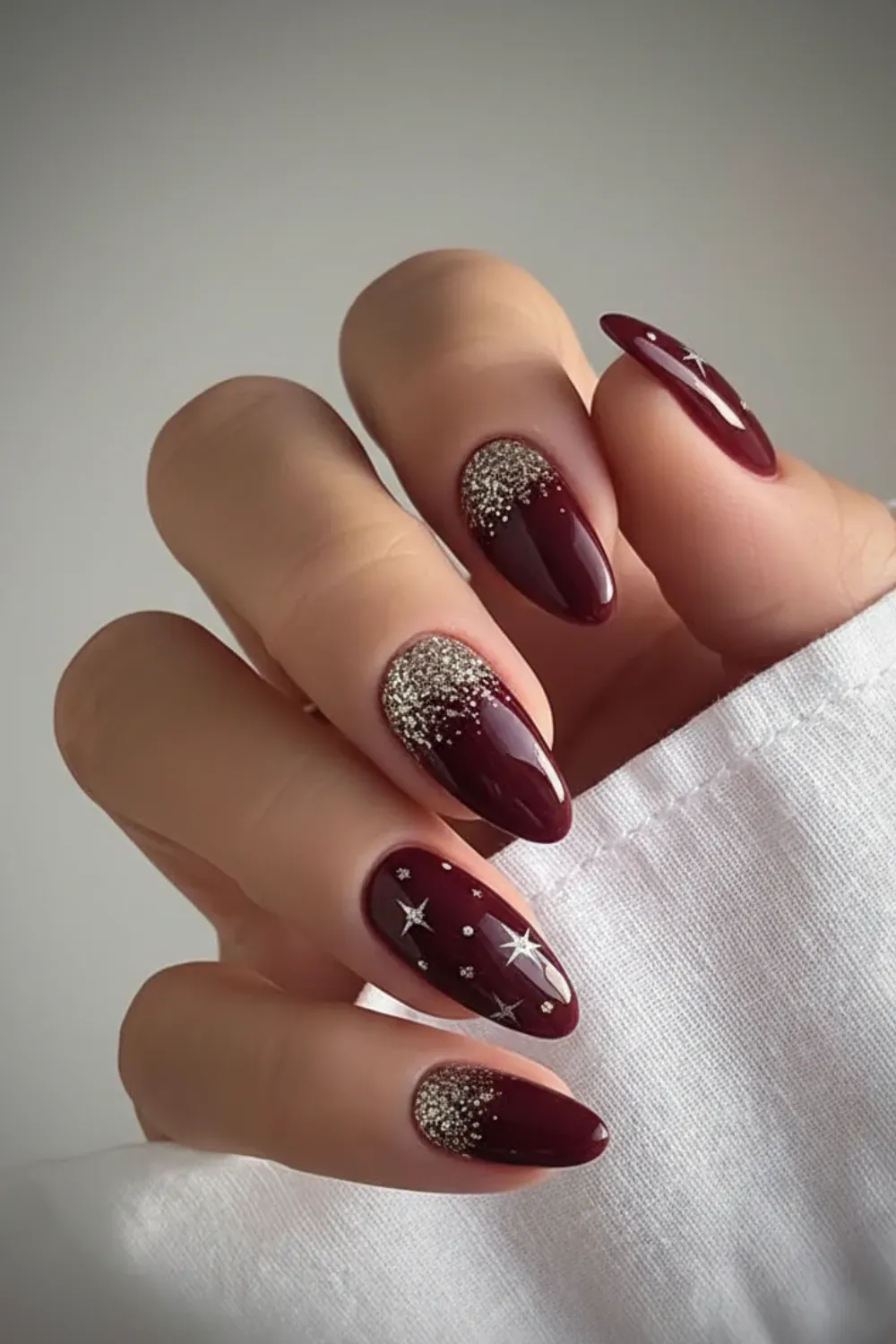 Hand with glossy almond-shaped burgundy nails, some featuring silver glitter at the cuticle and others adorned with silver stars.