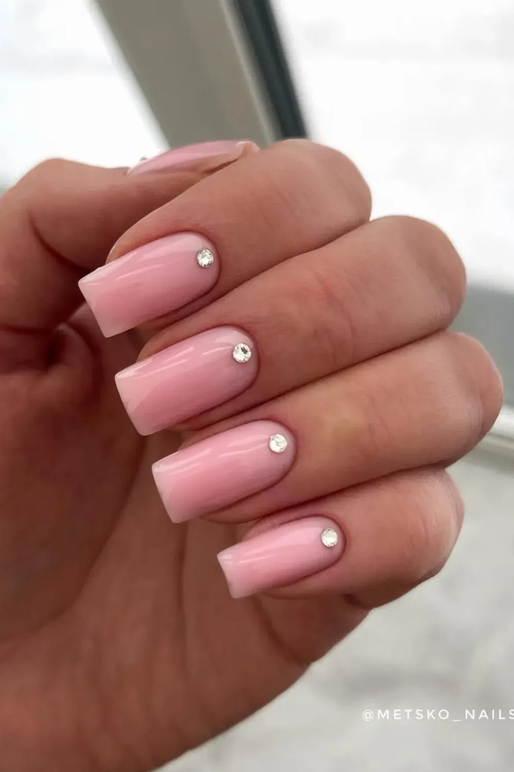 Hand with short square glossy light pink nails, each with a single clear rhinestone near the cuticle.