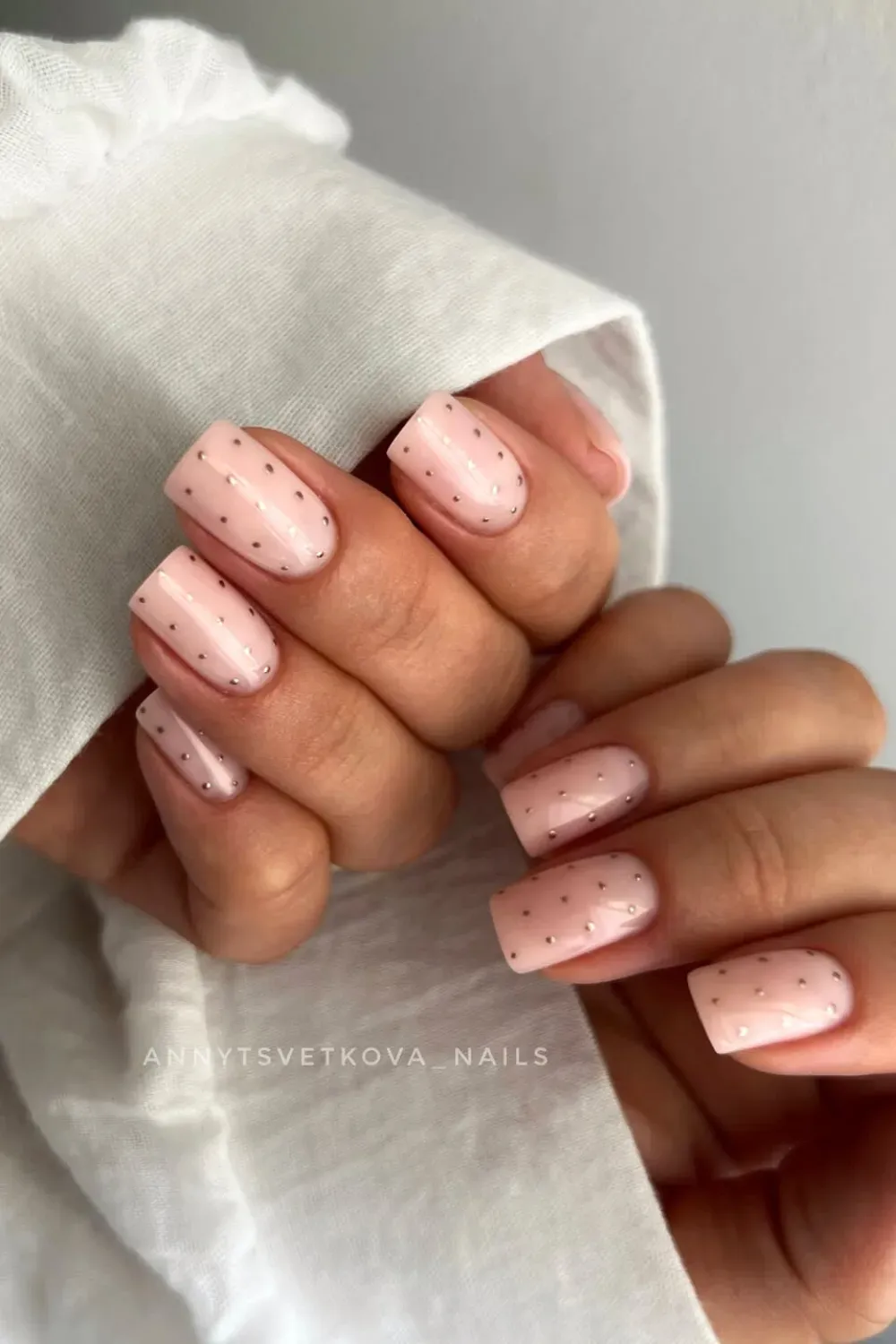 Hands with short square light pink nude nails with small gold glitter dots.