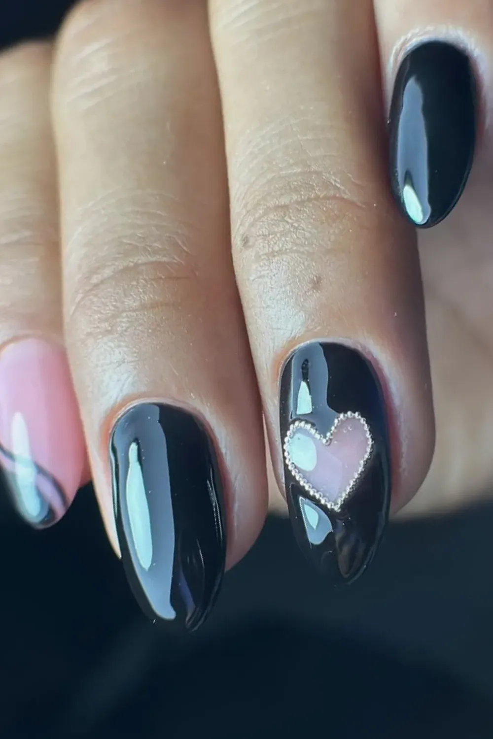 Oval nails featuring solid glossy black on most fingers, and an accent nail with a glossy black base and a large, pink raised heart charm.