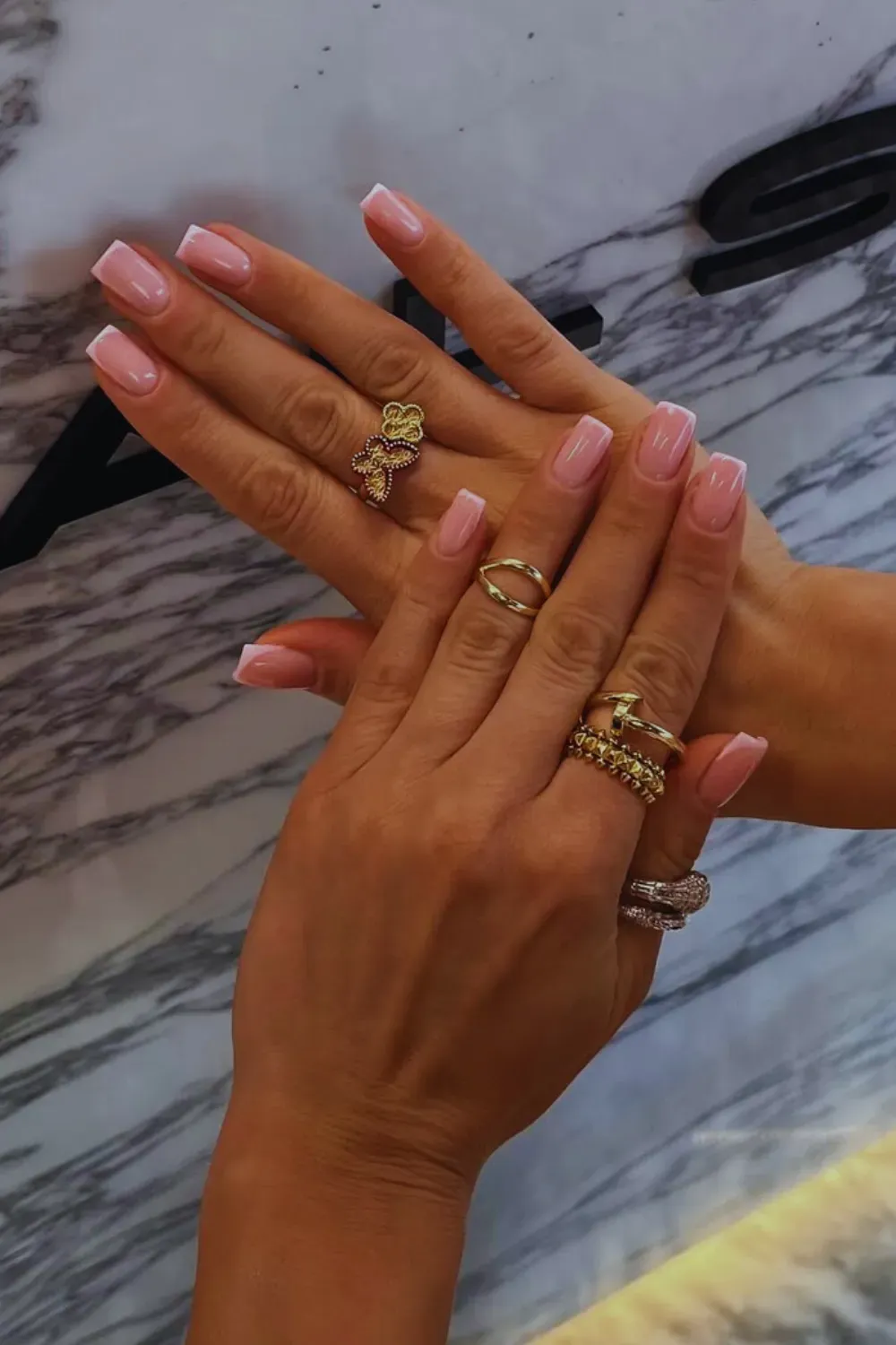 Elegant short square nails with a soft pink base and thin white French tips, accessorized with gold rings and bracelets.