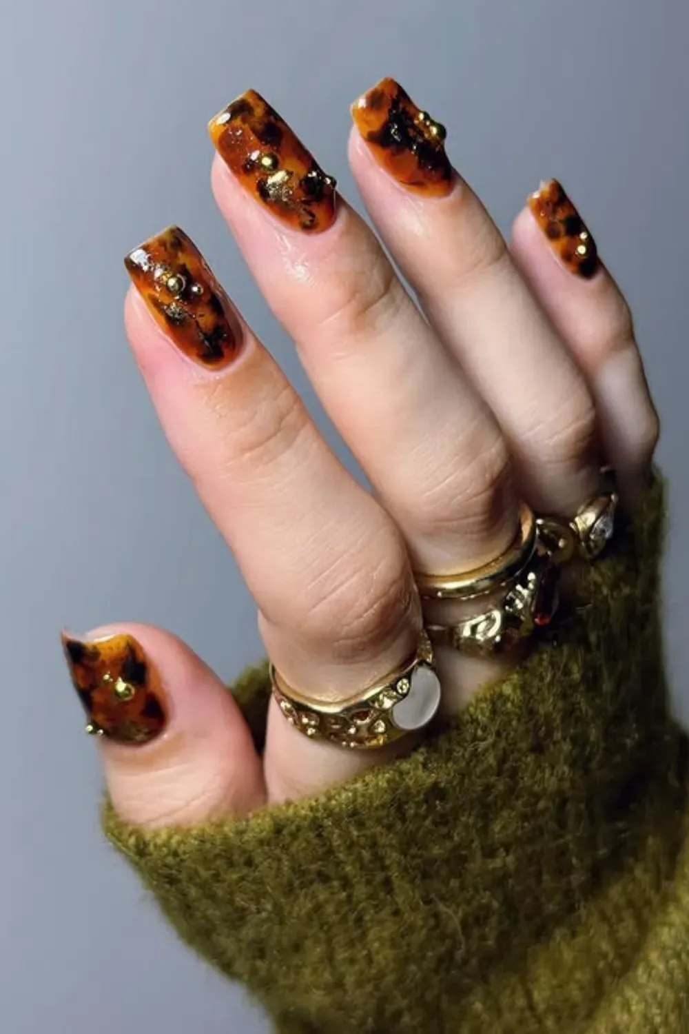 Hand with medium square tortoiseshell pattern nails adorned with small gold studs, wearing gold rings.