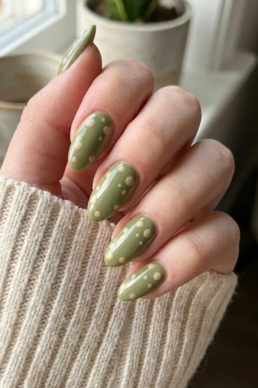 Olive Green and Nude Polka Dot Nails - Hand in a beige sweater sleeve showing almond nails in an earthy olive green shade featuring contrasting nude polka dots.