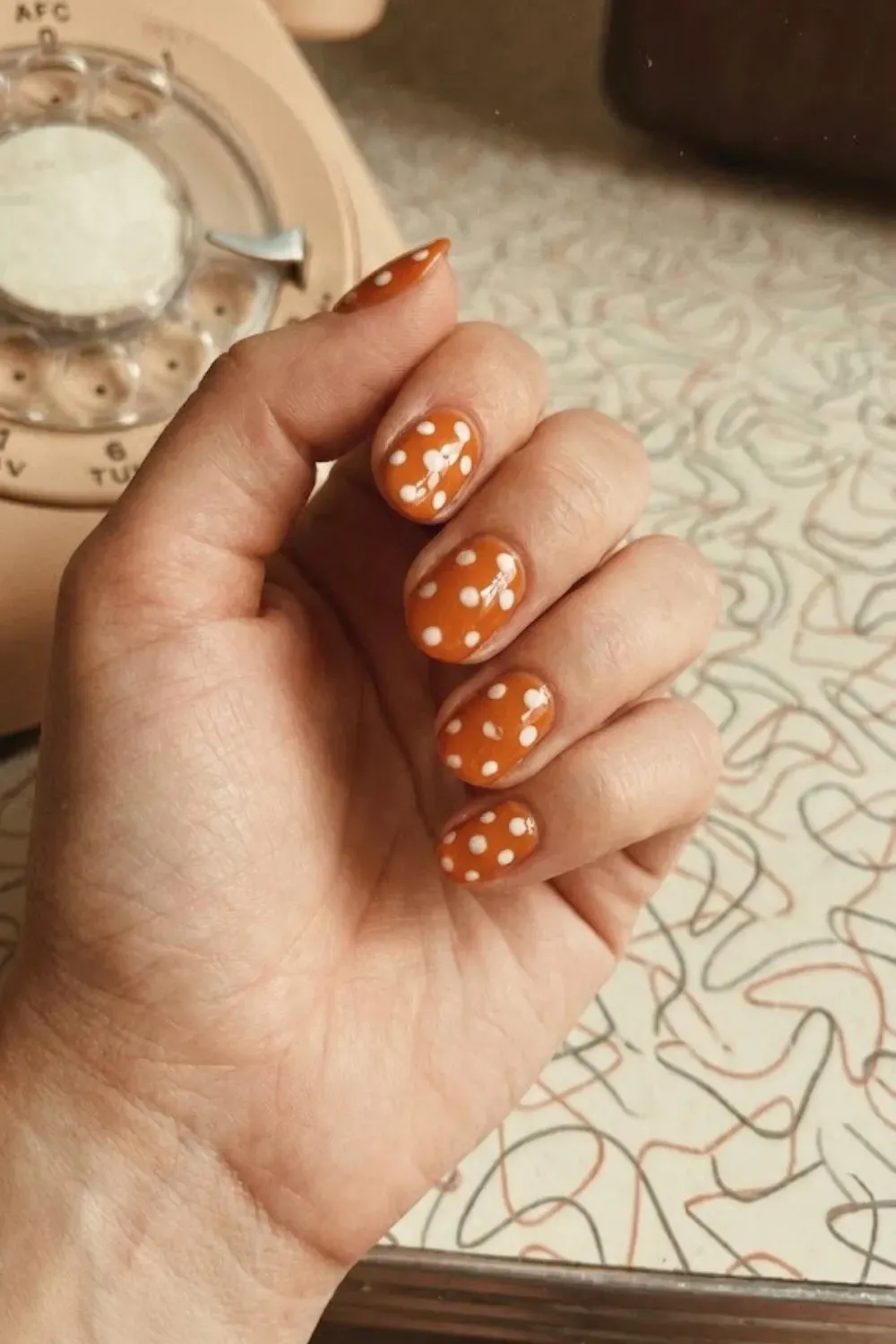 Retro Burnt Orange Polka Dot Nails - Short oval nails in a warm, retro burnt orange shade with white polka dots against a vintage background.