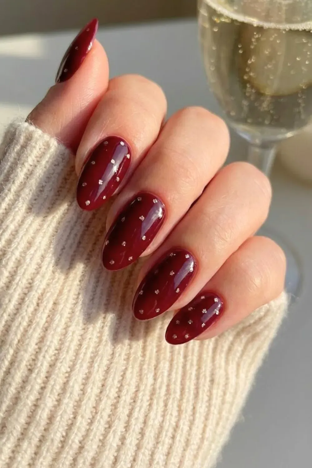 Burgundy and Silver Glitter Polka Dot Nails - Dark burgundy almond nails with subtle, sparkling silver glitter polka dots holding a champagne glass.
