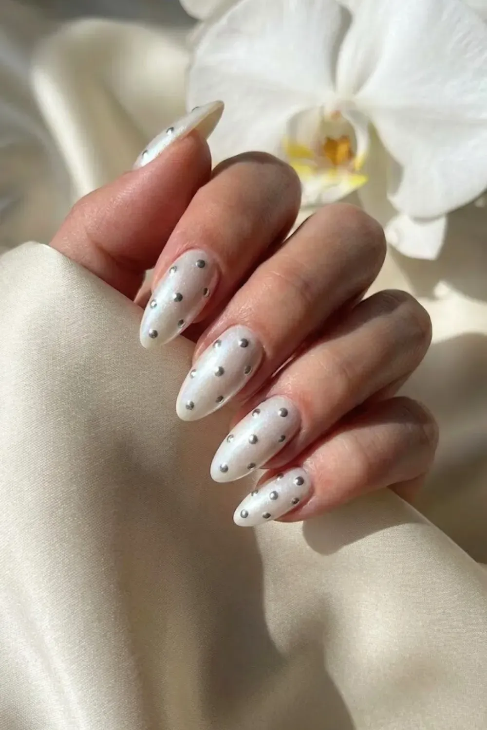 Pearlescent White and Silver Stud Nails - Shimmering pearl white almond nails adorned with raised metallic silver polka dot studs near a white orchid.
