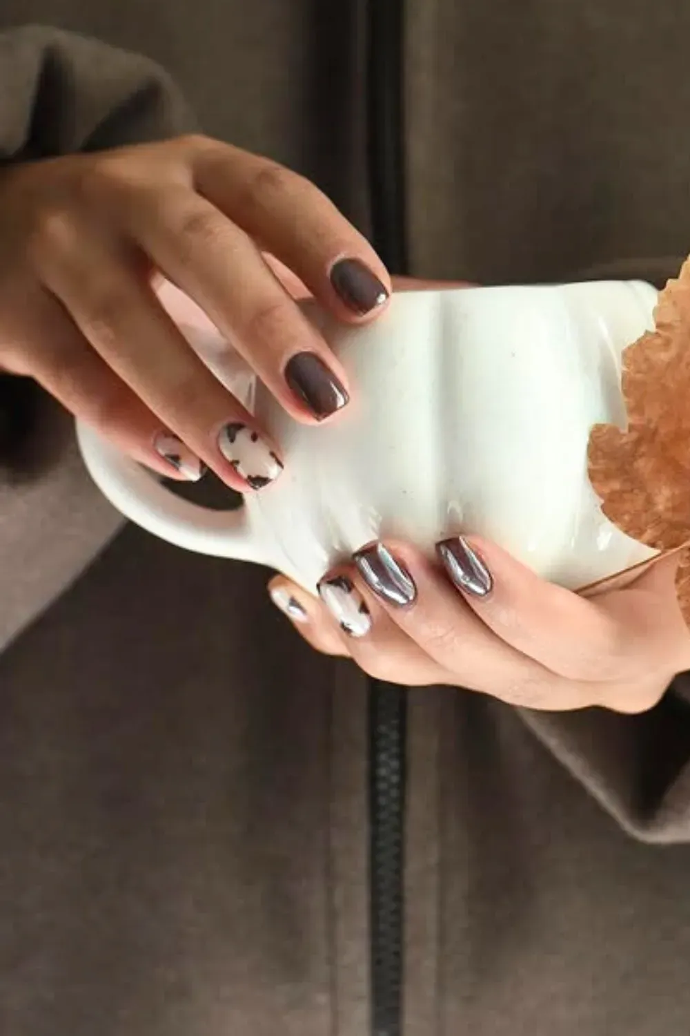 Cozy Espresso and Speckled Accent Nails Short, rounded nails in a glossy dark brown, with an accent nail featuring a white base and a small, speckled brown/black pattern. Hand holds a white mug and a dried maple leaf.
