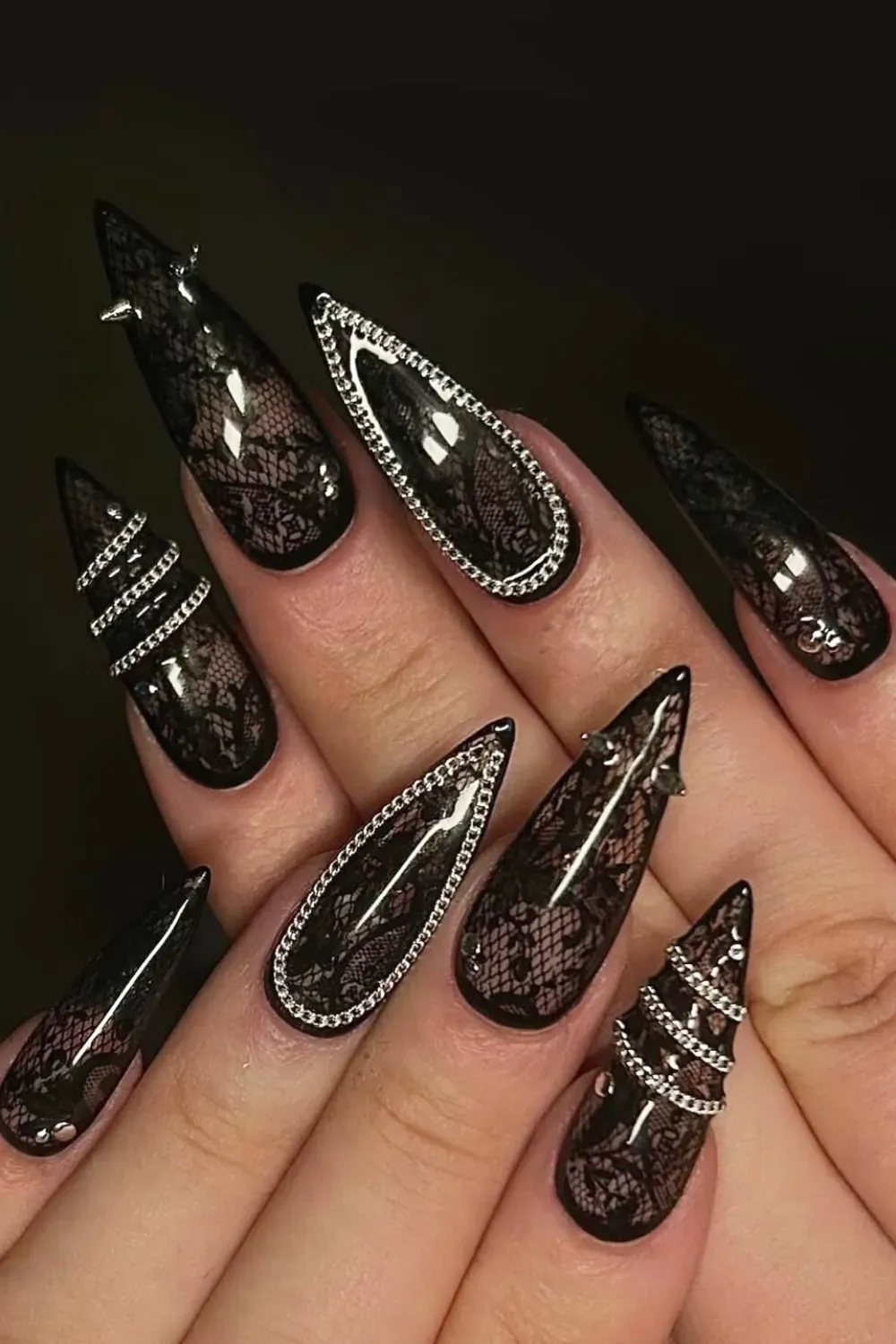 Sharp stiletto nails in glossy black with transparent black lace pattern, silver chains, and metallic spikes.