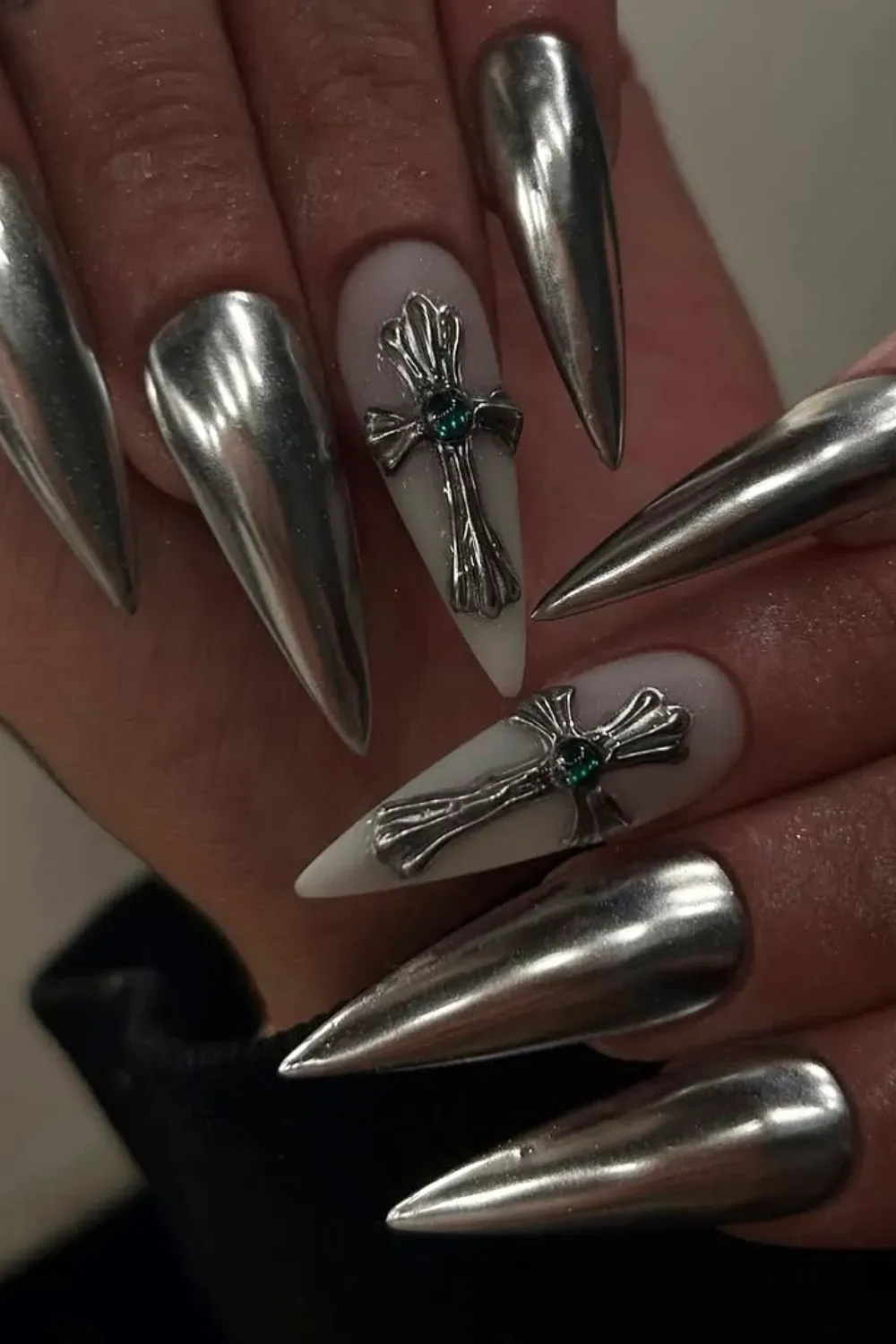 Sharp stiletto nails fully covered in liquid silver chrome, with two white accent nails featuring 3D silver chrome crosses with green stones.