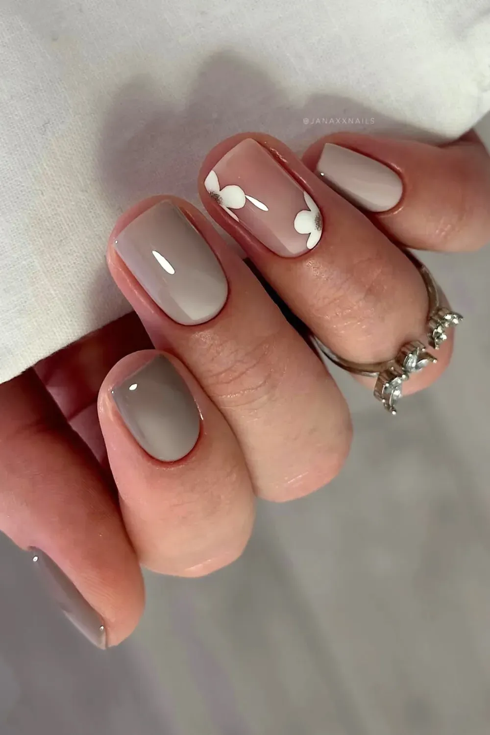 Greige and Delicate Floral Nails	Hand showing short, rounded nails in taupe/greige with one accent nail featuring small white flower decals on a nude base.