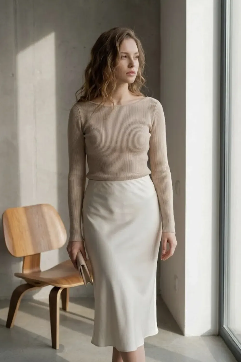 Beige Ribbed Top & Silk Slip Skirt – Woman standing in soft natural light wearing a fitted beige ribbed long-sleeve top tucked into a flowing cream silk slip skirt, holding a neutral clutch.