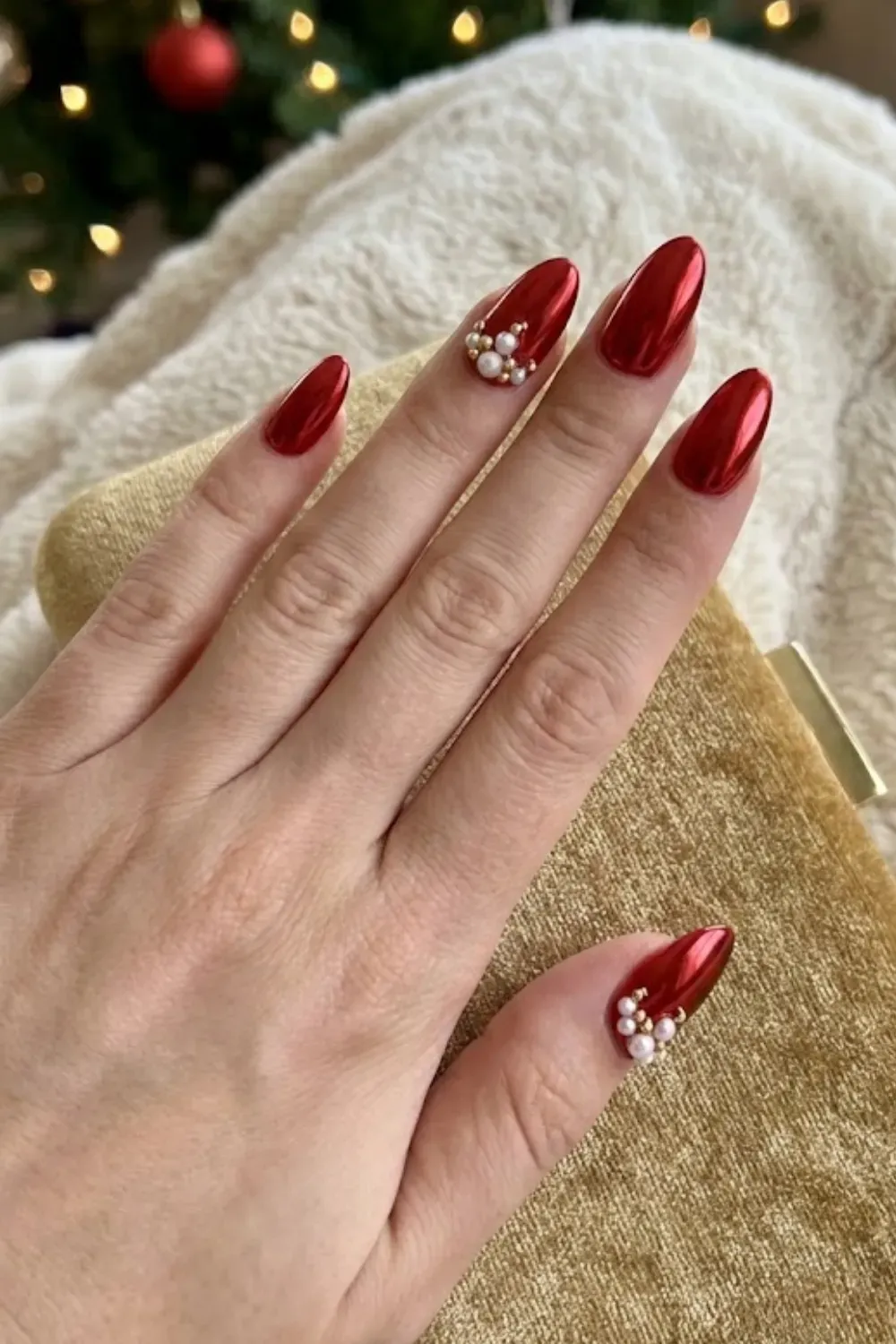 Red Chrome Nails with Pearl Accents - Hand holding a gold clutch featuring metallic red nails adorned with clusters of pearls and gold beads.
