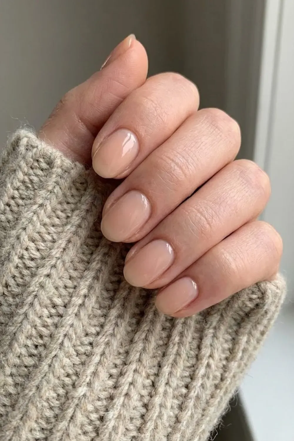 Sheer Glossy Nude Oval - Short natural oval nails with a high-shine sheer nude polish, paired with a beige knit sleeve for a cozy minimalist look.