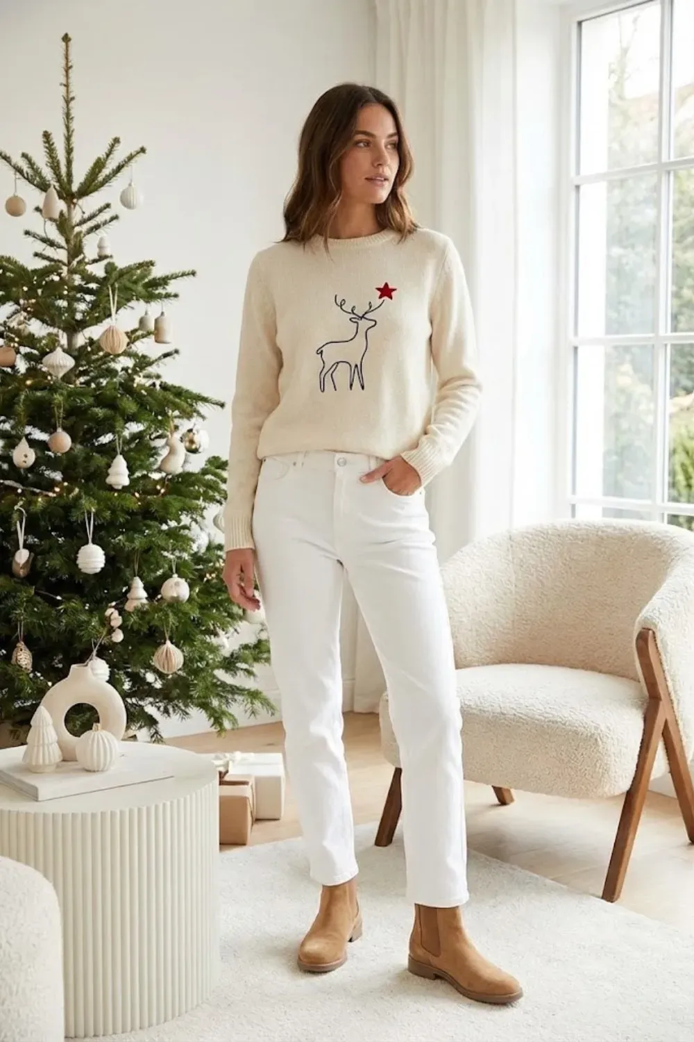 Woman standing by christmas tree wearing cream sweater with minimal reindeer design and white jeans