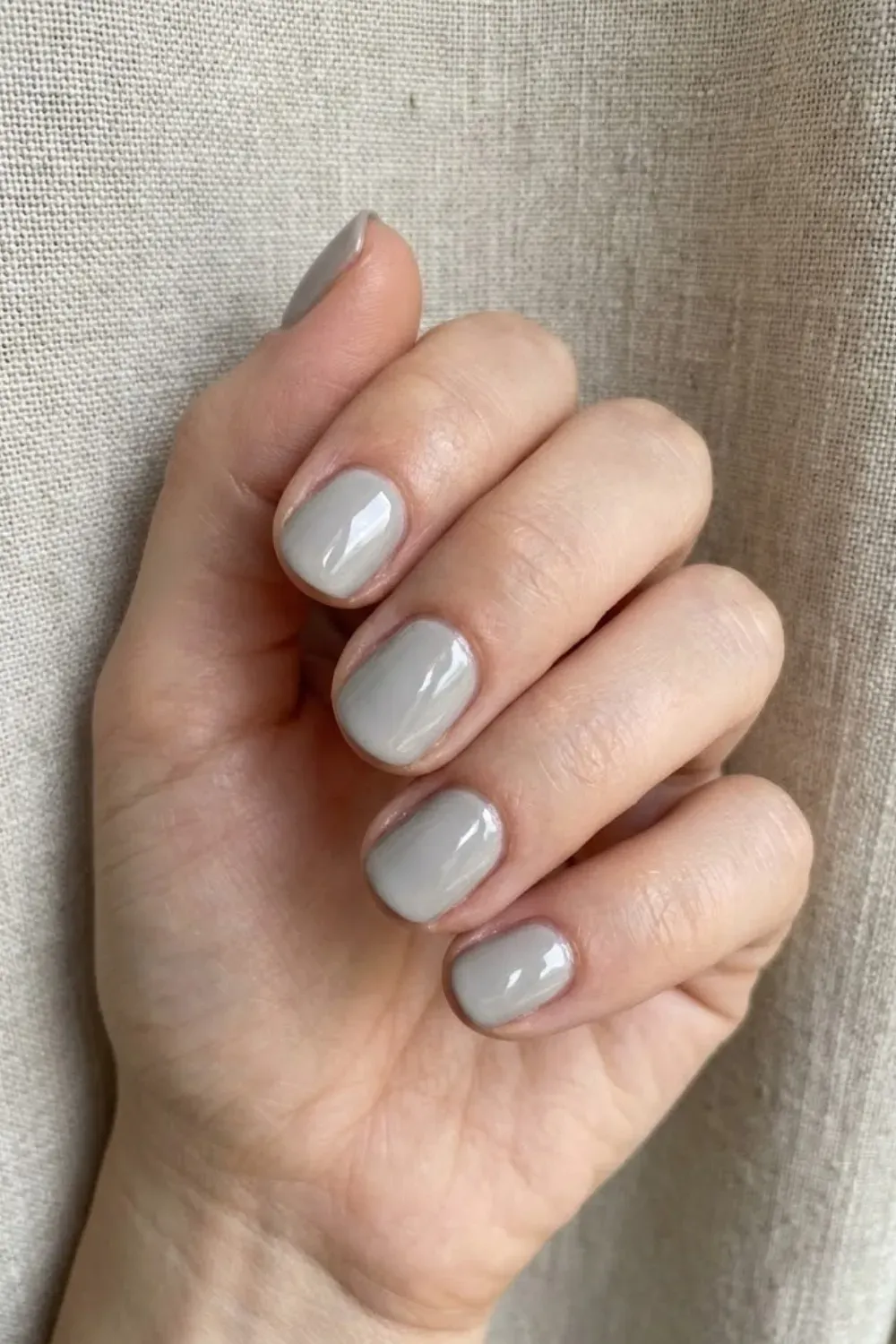 Soft Putty Gray Round - Short round nails painted in a light glossy putty gray polish, contrasting with a neutral fabric background.