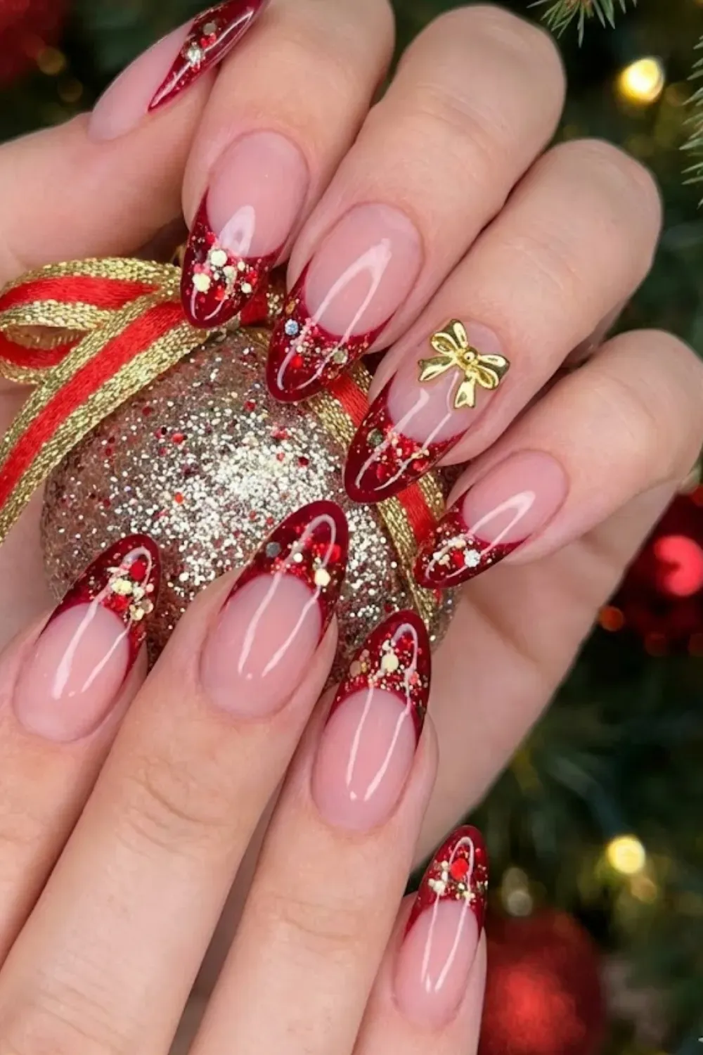 Ruby Glitter Gold Bow Nails — almond nails with red glitter tips and a 3D gold ribbon charm at the cuticle.