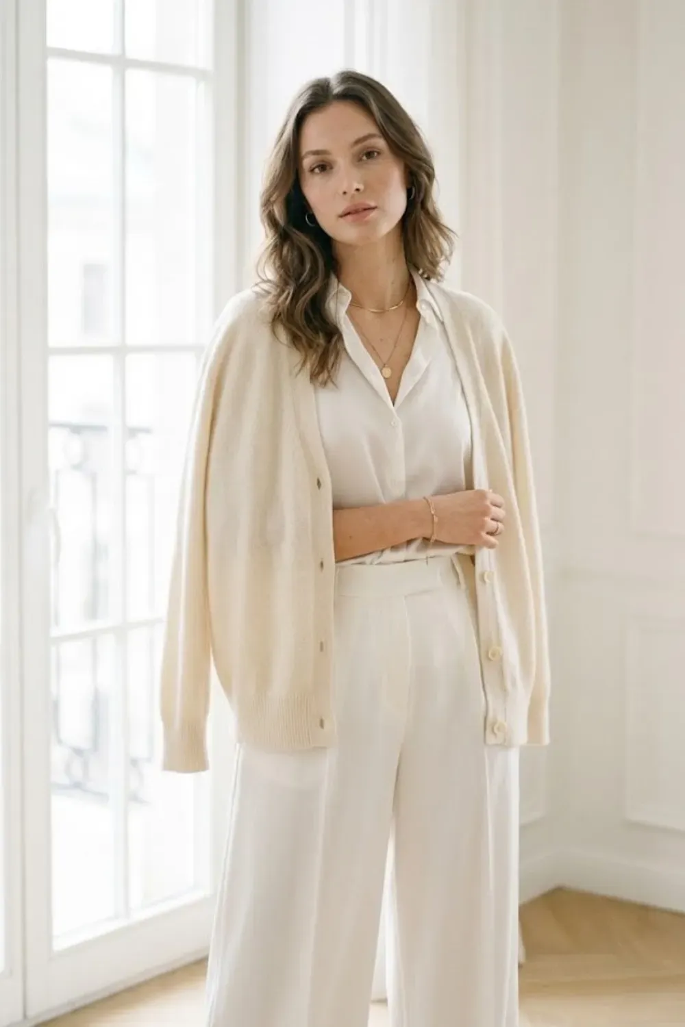 White Monochromatic Cardigan & Wide Leg Pants – All-white minimalist outfit featuring a cream cardigan worn over a silky white blouse and white wide-leg trousers, standing in a bright room.