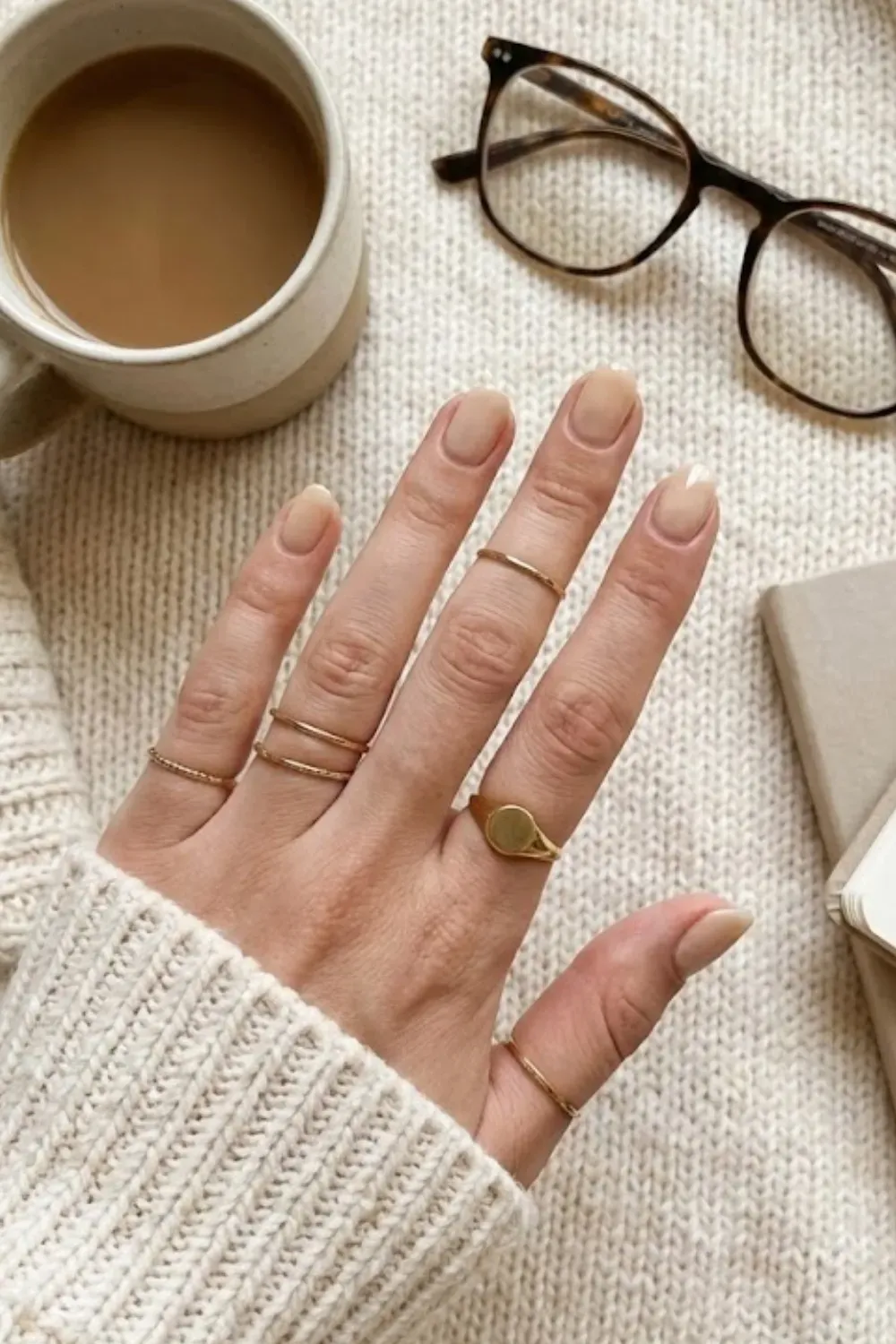 Minimal Sheer Nude Round Nails – Short round nails with a sheer nude wash, styled with multiple gold rings and a chunky knit sweater.