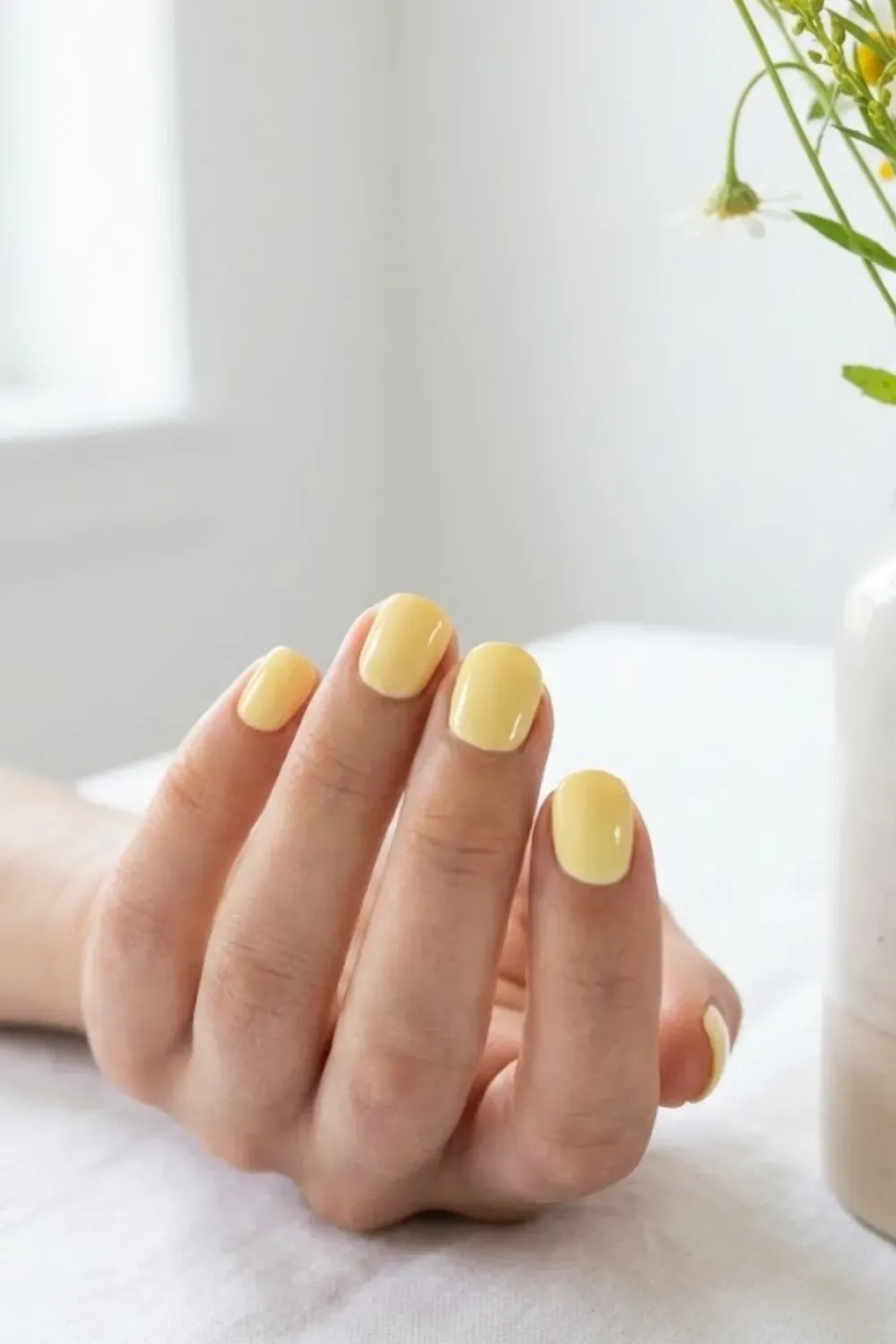 Short square nails painted in a soft pastel butter yellow color, holding a vase of wildflowers.