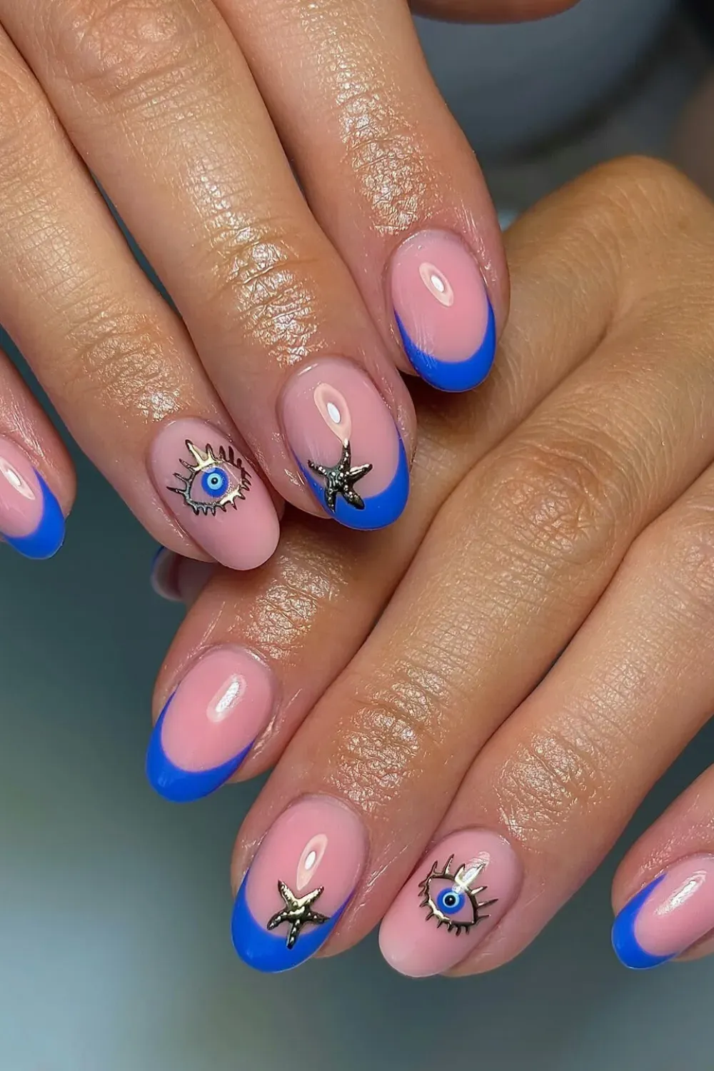 Summer Evil Eye French Tips – Oval nails with bright blue French tips on a nude base, decorated with gold evil eye and starfish 3D charms.
