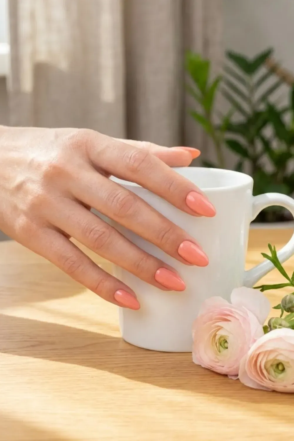Bright coral pink nails on almond shape, holding a white coffee mug.