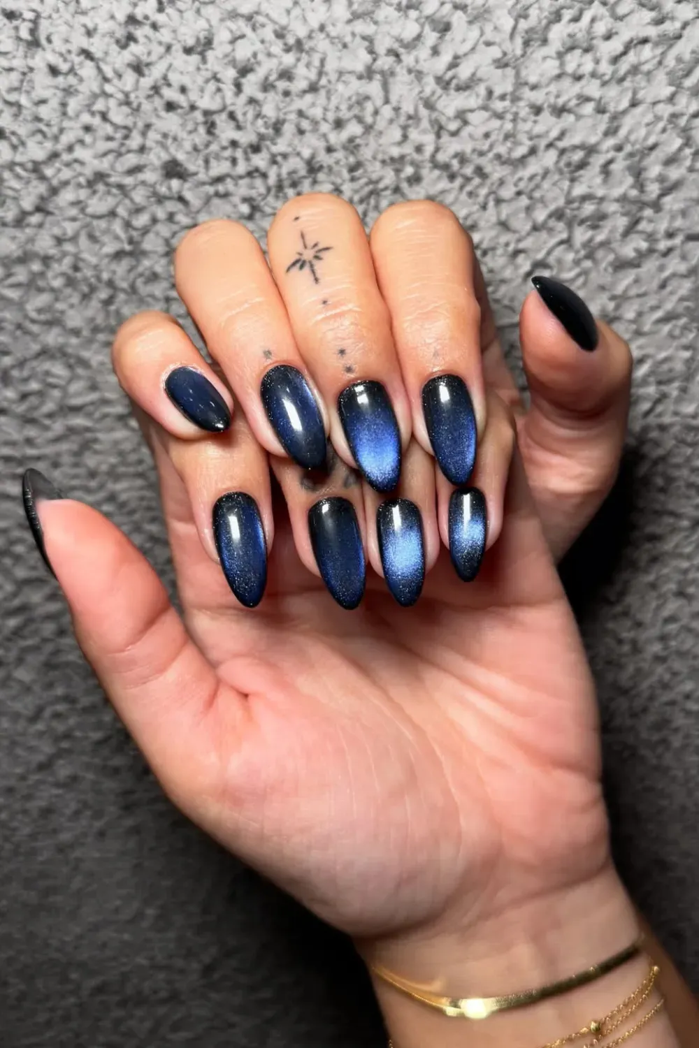 Galaxy Cat Eye Stiletto Nails – Dark navy magnetic cat eye nails with shimmering galactic blue effects on an almond-stiletto shape, paired with minimalist finger tattoos.