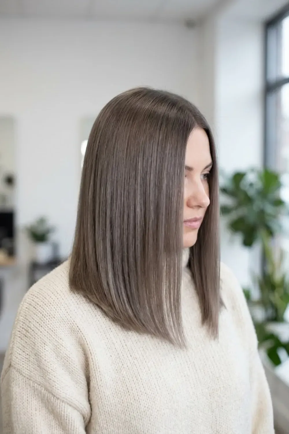 Sleek Ash Mushroom Brown Lob – Side profile of a woman with a sleek, straight ash mushroom brown long bob haircut indoors.