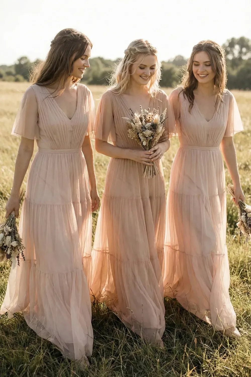 Three bridesmaids walking through a tall grass field wearing dusty rose tulle dresses with tiered skirts, flutter sleeves, and pleated V-necks, holding dried flower bouquets.
