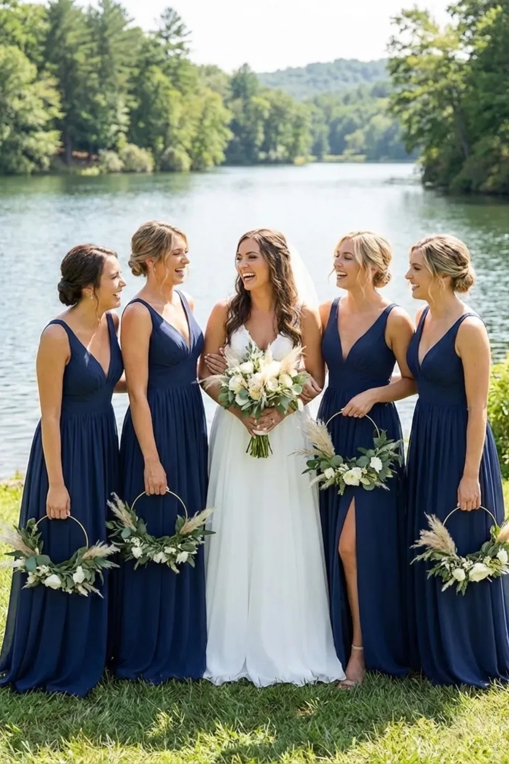 Bridesmaids in floor-length navy blue V-neck chiffon dresses standing on grass by a lake, holding unique gold hoop bouquets with white flowers and greenery.