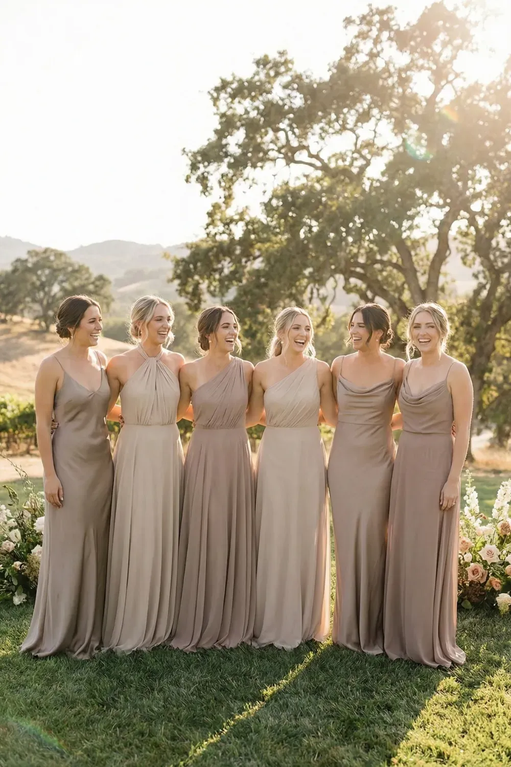 A group of six bridesmaids laughing in a vineyard wearing mismatched floor-length silk dresses in varying shades of taupe and champagne, featuring halter, one-shoulder, and cowl necklines.