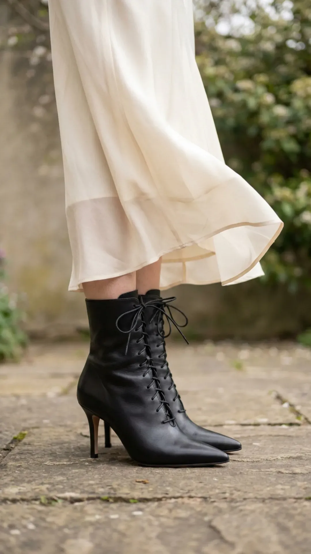 Close-up detail of black pointed-toe Victorian-style lace-up stiletto boots paired with a flowing cream silk midi skirt.