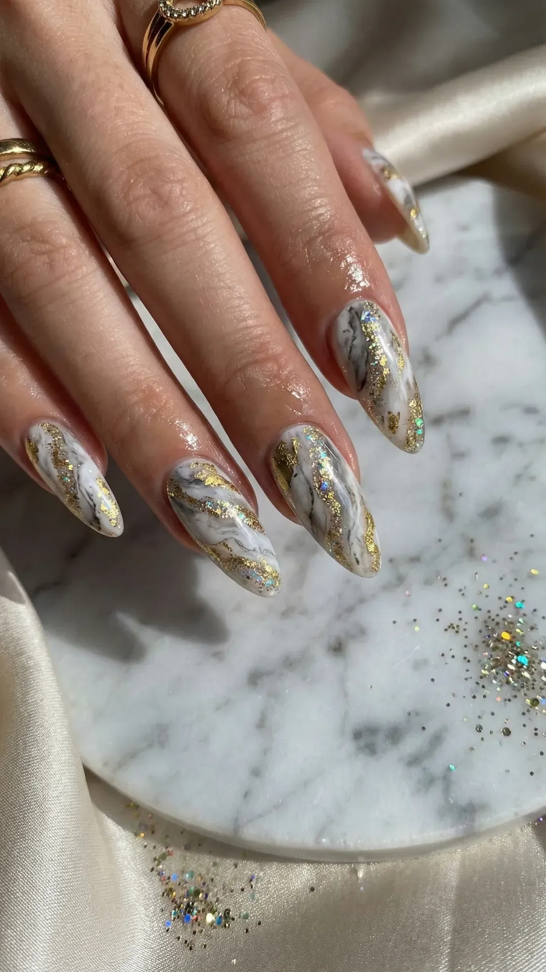 Almond nails with white marble design and gold glitter veins on marble tray background