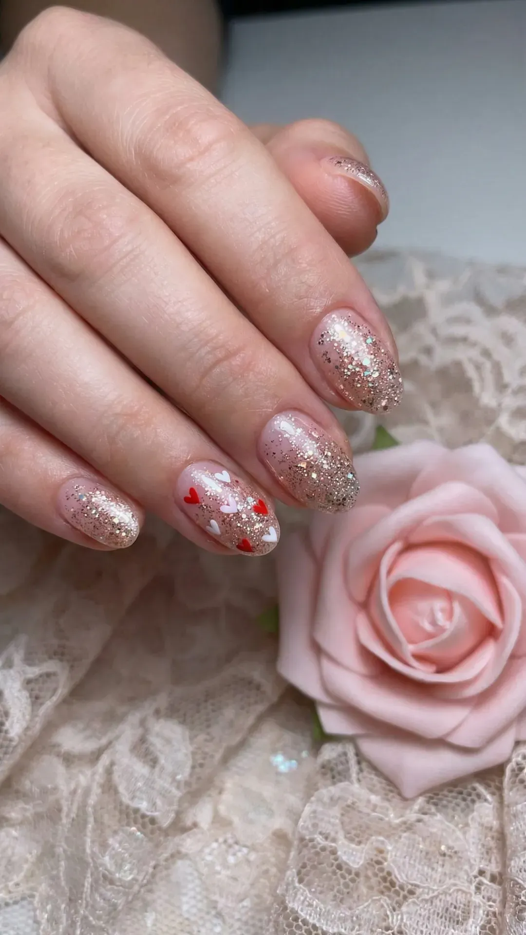 Short almond nails with nude glitter base and tiny red heart accents beside pink rose