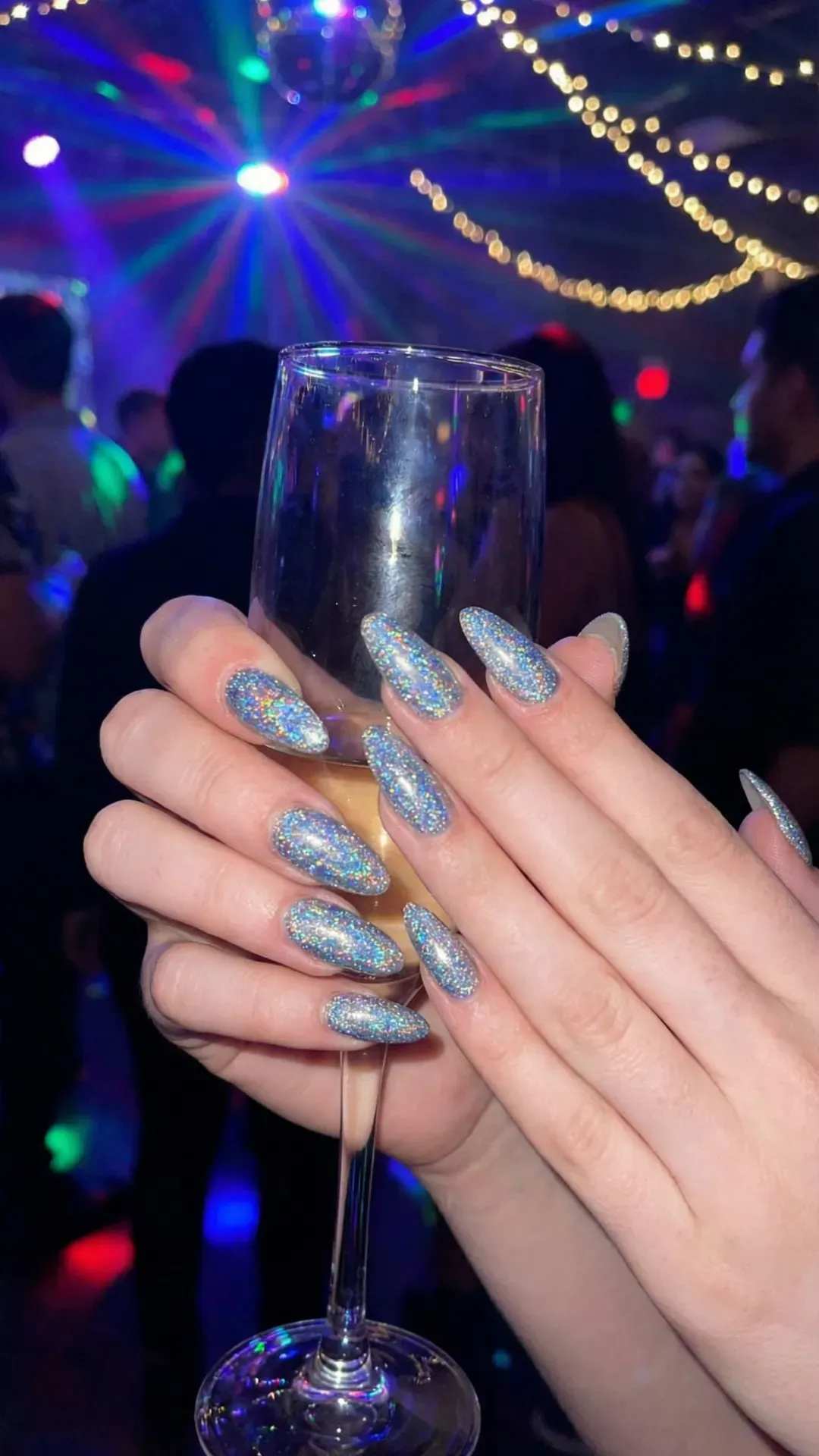 Long almond nails with holographic blue glitter holding champagne glass, colorful party lights background