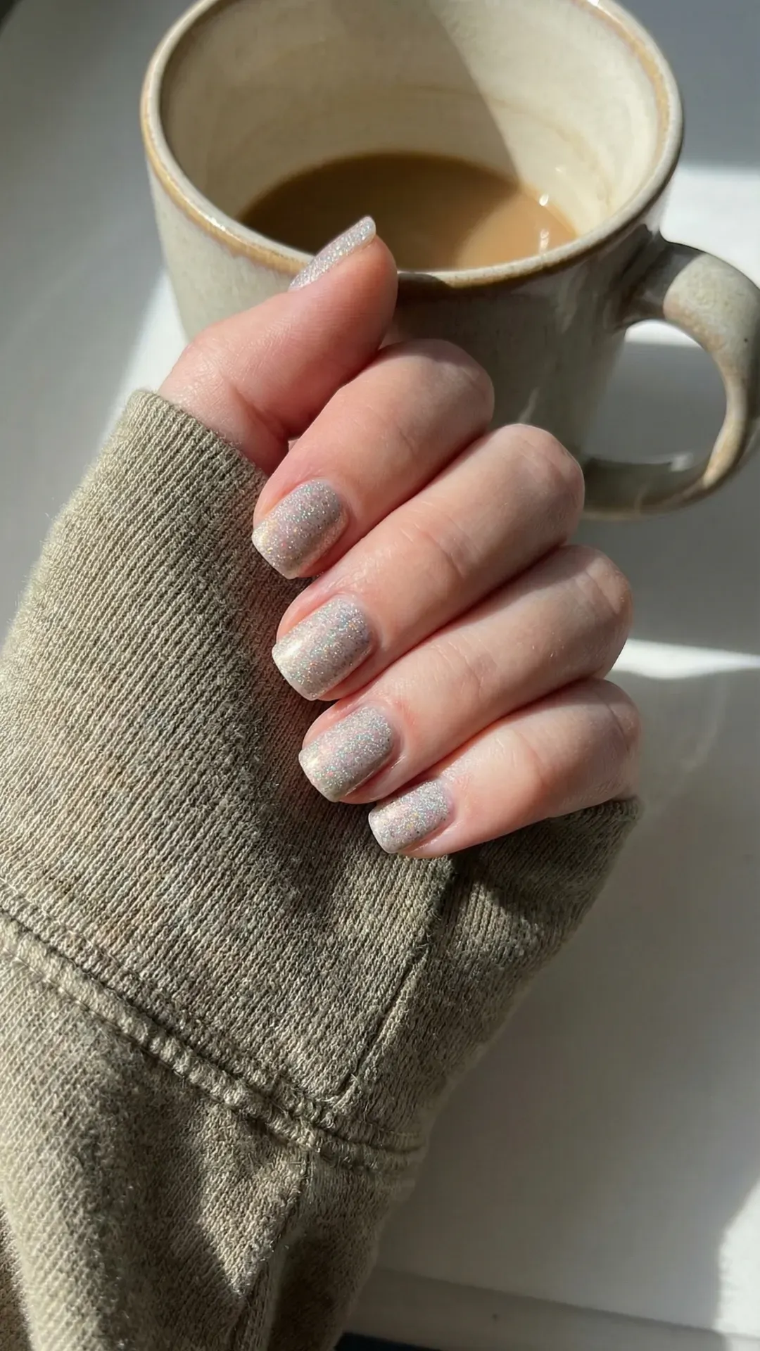 Short natural nails with soft silver micro-glitter polish holding coffee mug, sunlight window aesthetic