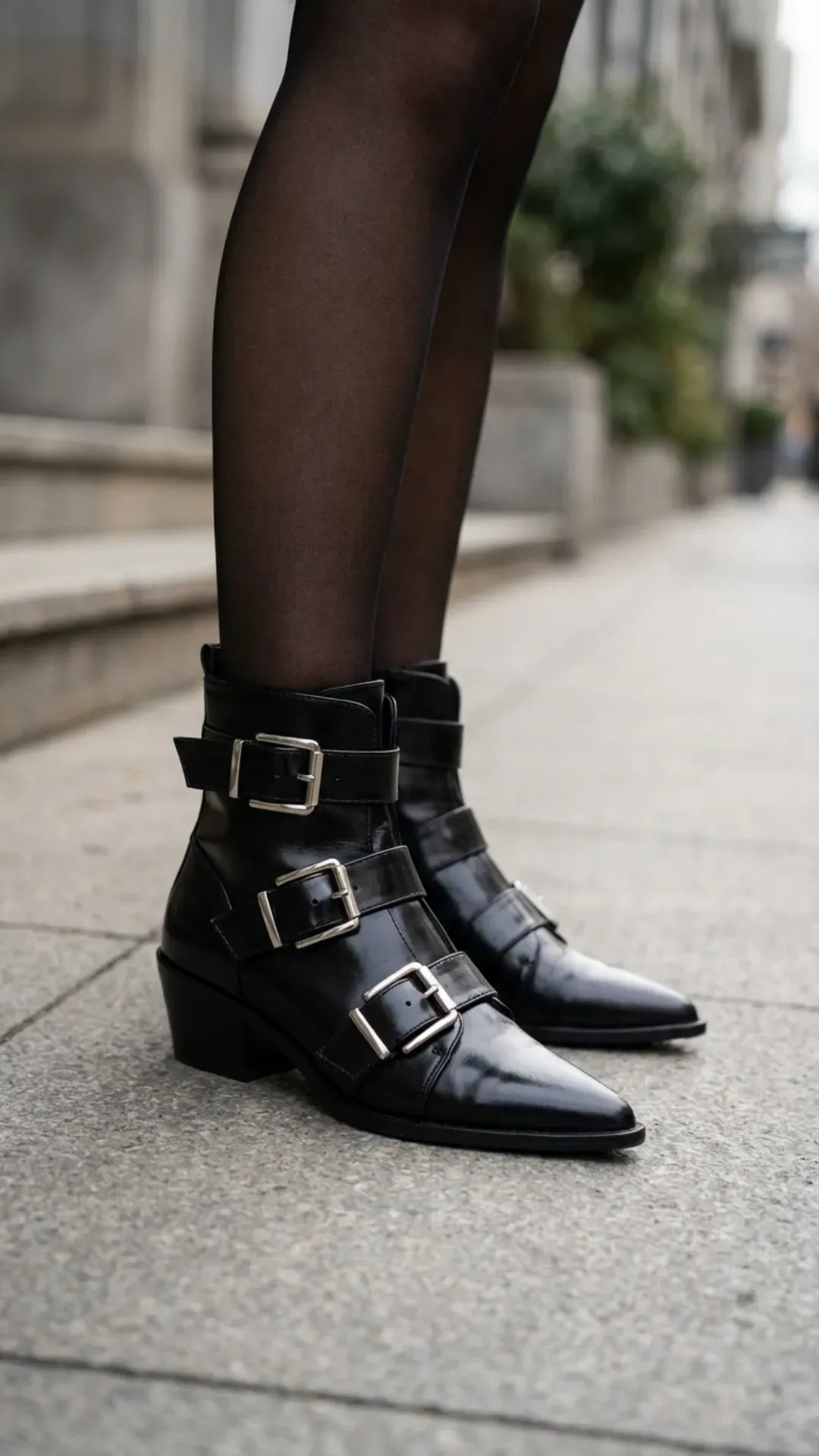 Detailed shot of black leather pointed-toe boots featuring multiple silver buckles, worn with sheer black tights on stone steps.