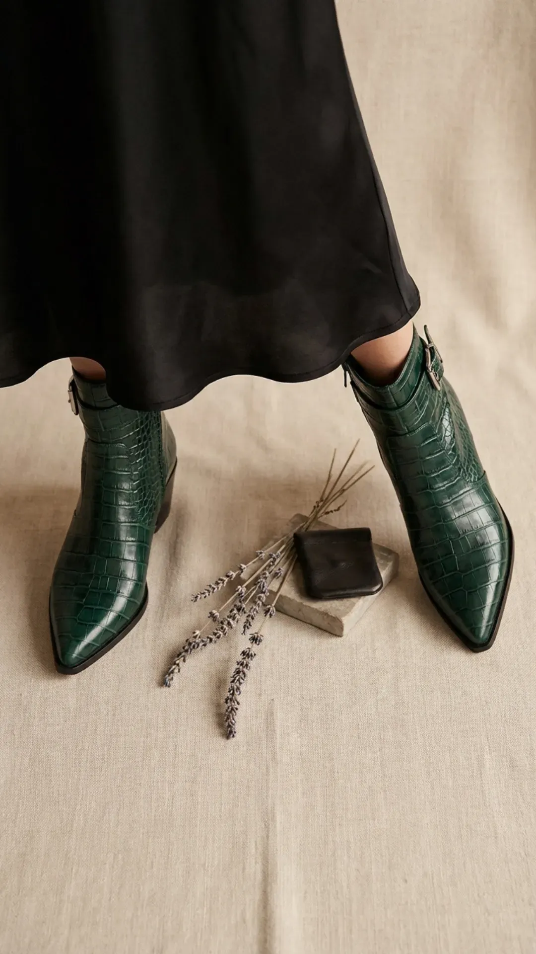 Top-down view of emerald green mock-croc leather pointed ankle boots styled with a black satin midi skirt and dried lavender sprigs.
