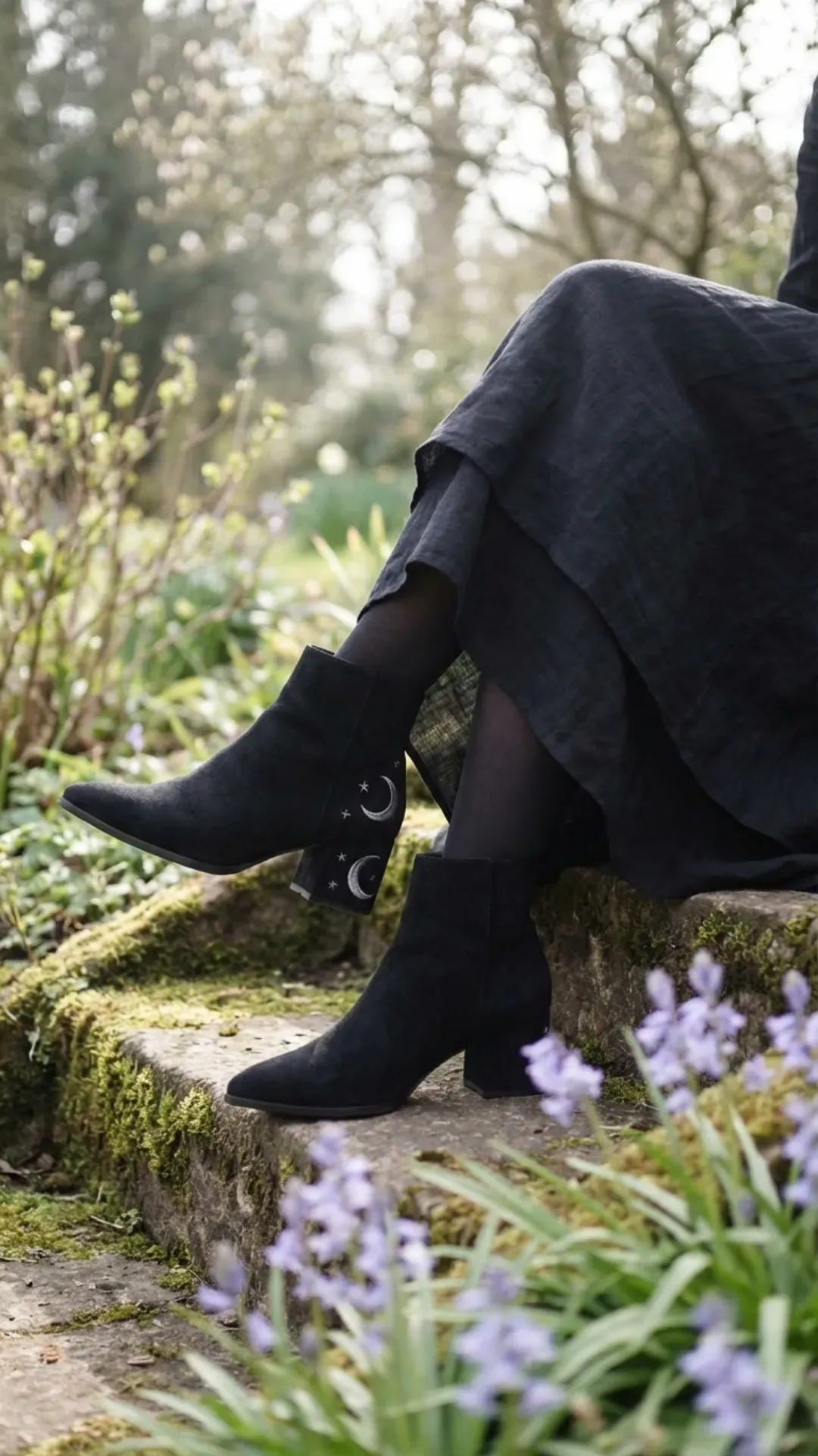 Black suede ankle boots featuring intricate silver moon and star celestial embroidery on the heel, resting on mossy stone steps near bluebells.