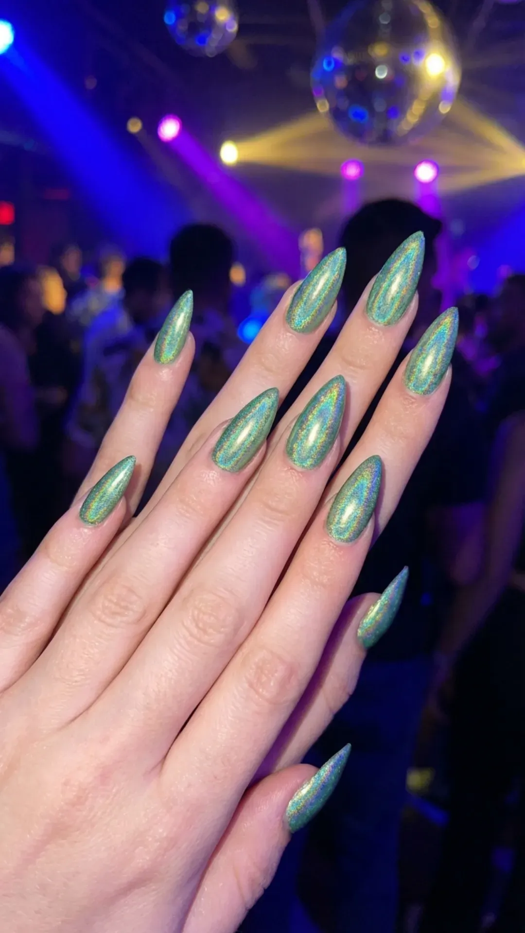 Holographic green chrome stiletto nails reflecting rainbow tones in club lighting
