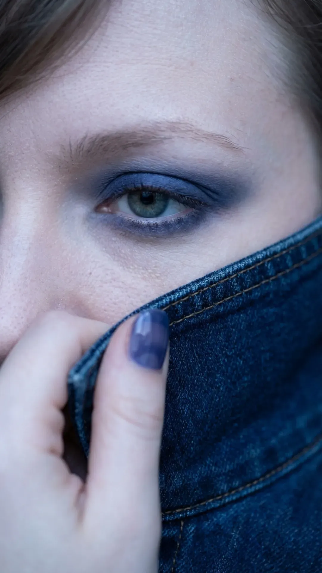 Close-up of a matte indigo blue smoky eye with feathered brows and sheer indigo jelly nails touching a denim collar.