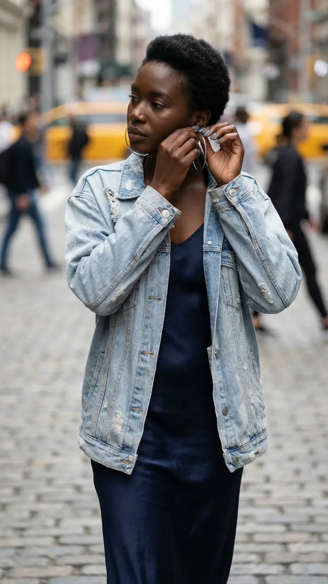 Stylish woman with deep skin tone wearing a light wash denim jacket, silver hoops, and a matte grayish-blue short square manicure.