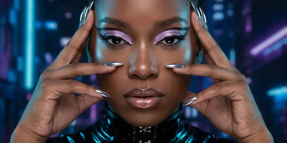 Woman with deep skin tone wearing Y3K cyber-luxe makeup with lilac holographic eyeshadow, glass skin, and liquid silver chrome stiletto nails.