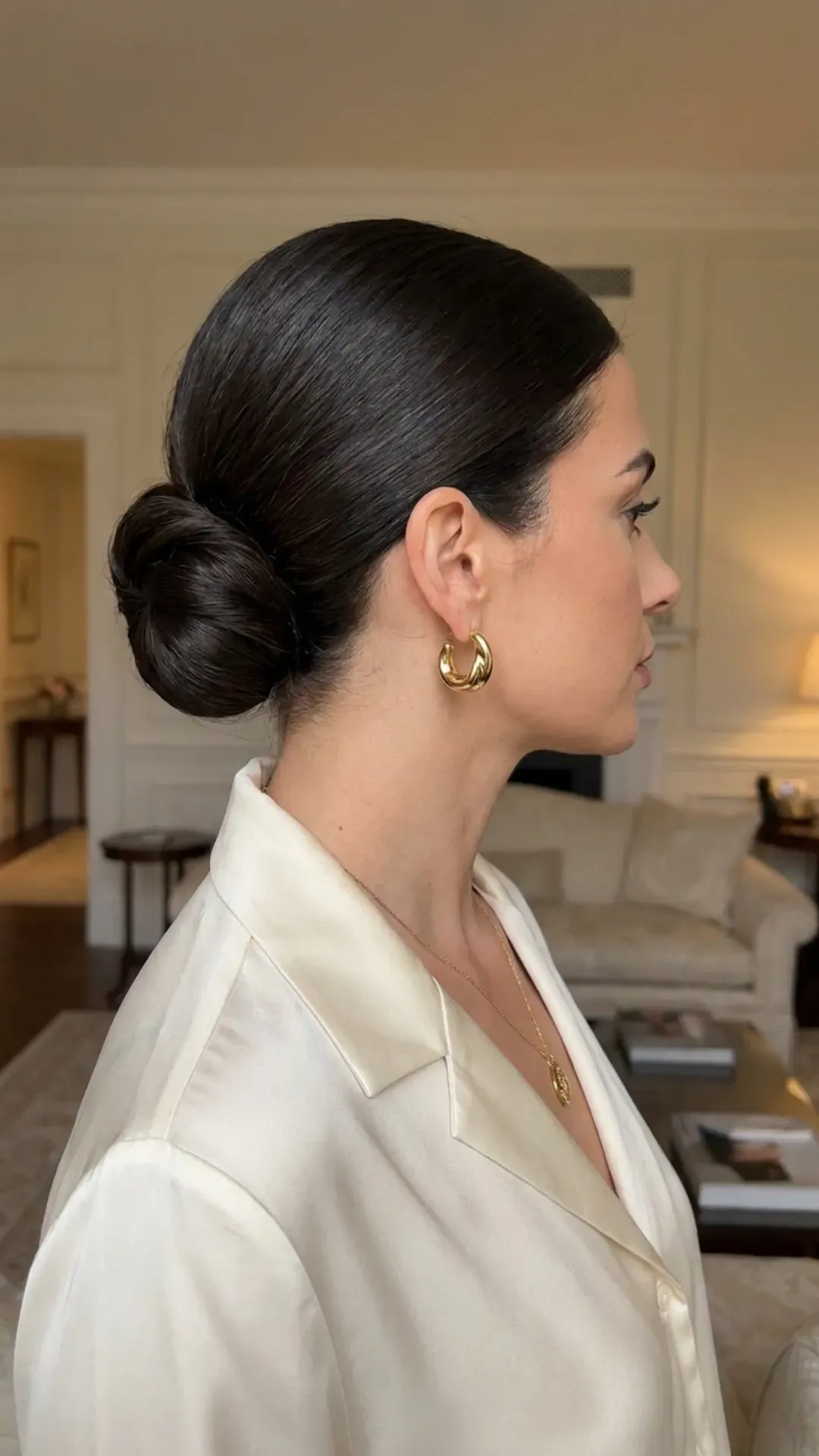 Woman with a sleek low chignon hairstyle, perfectly smoothed and styled, accessorized with a golden earring and necklace, exuding sophistication and luxury.