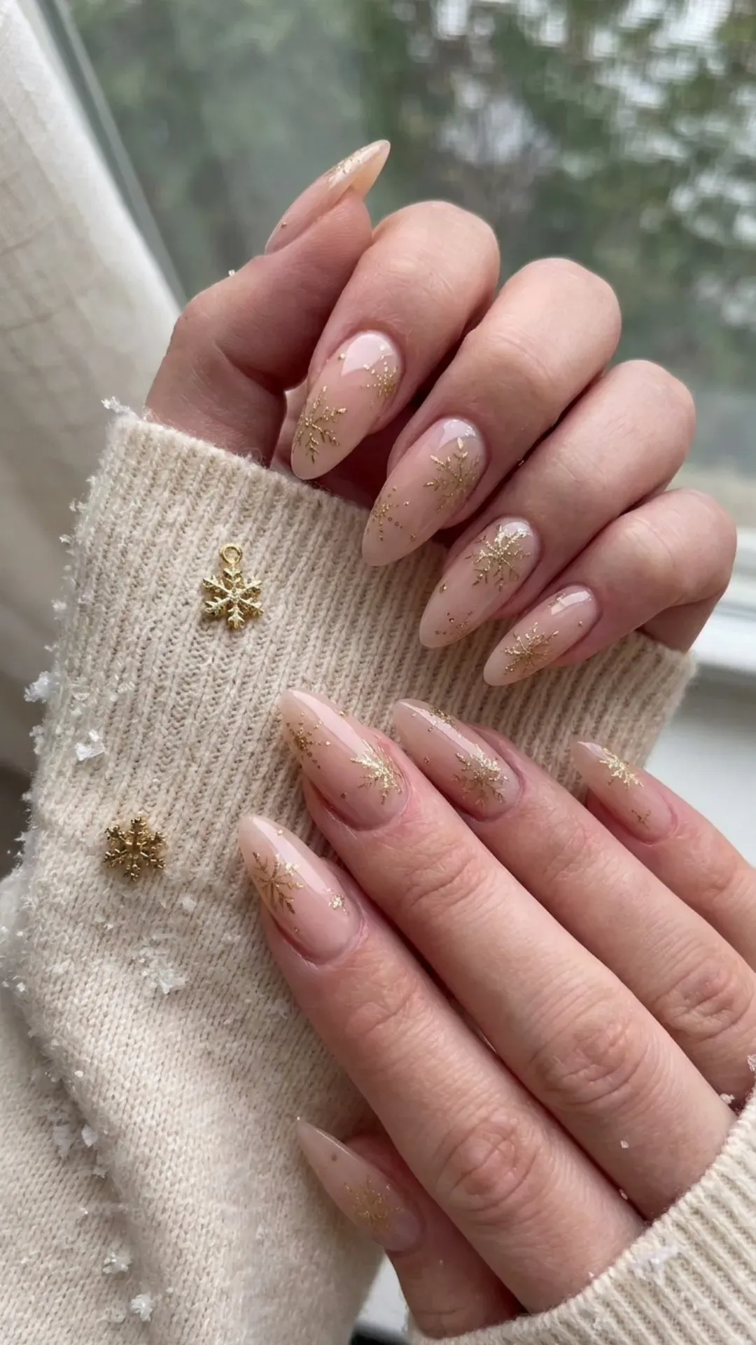 Elegant nude pink acrylic nails with a glossy finish and golden snowflake designs, perfect for a subtle winter wonderland theme.