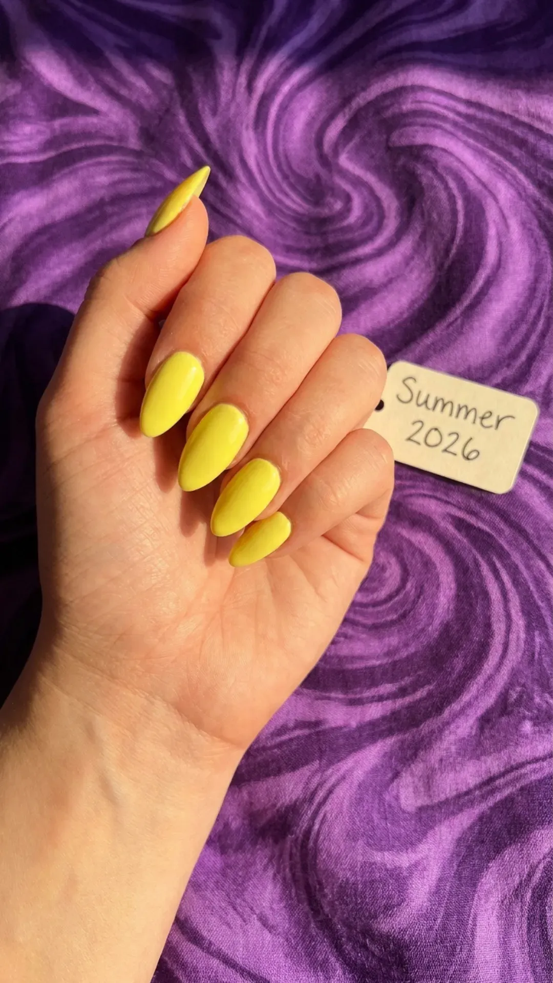 Bright yellow almond-shaped nails against a purple background, emphasizing a cheerful and bold nail color for summer 2026.