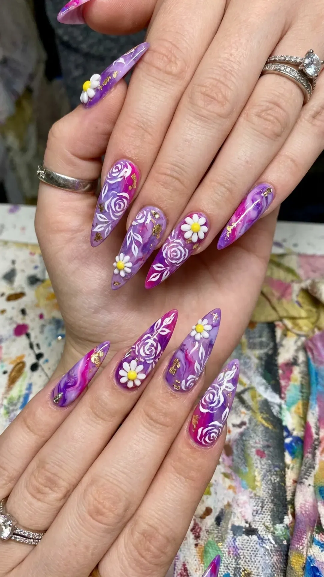 Stiletto nails featuring a swirling mix of purple and pink shades, complemented by white floral patterns and small daisy accents, creating a bold and artistic nail design.