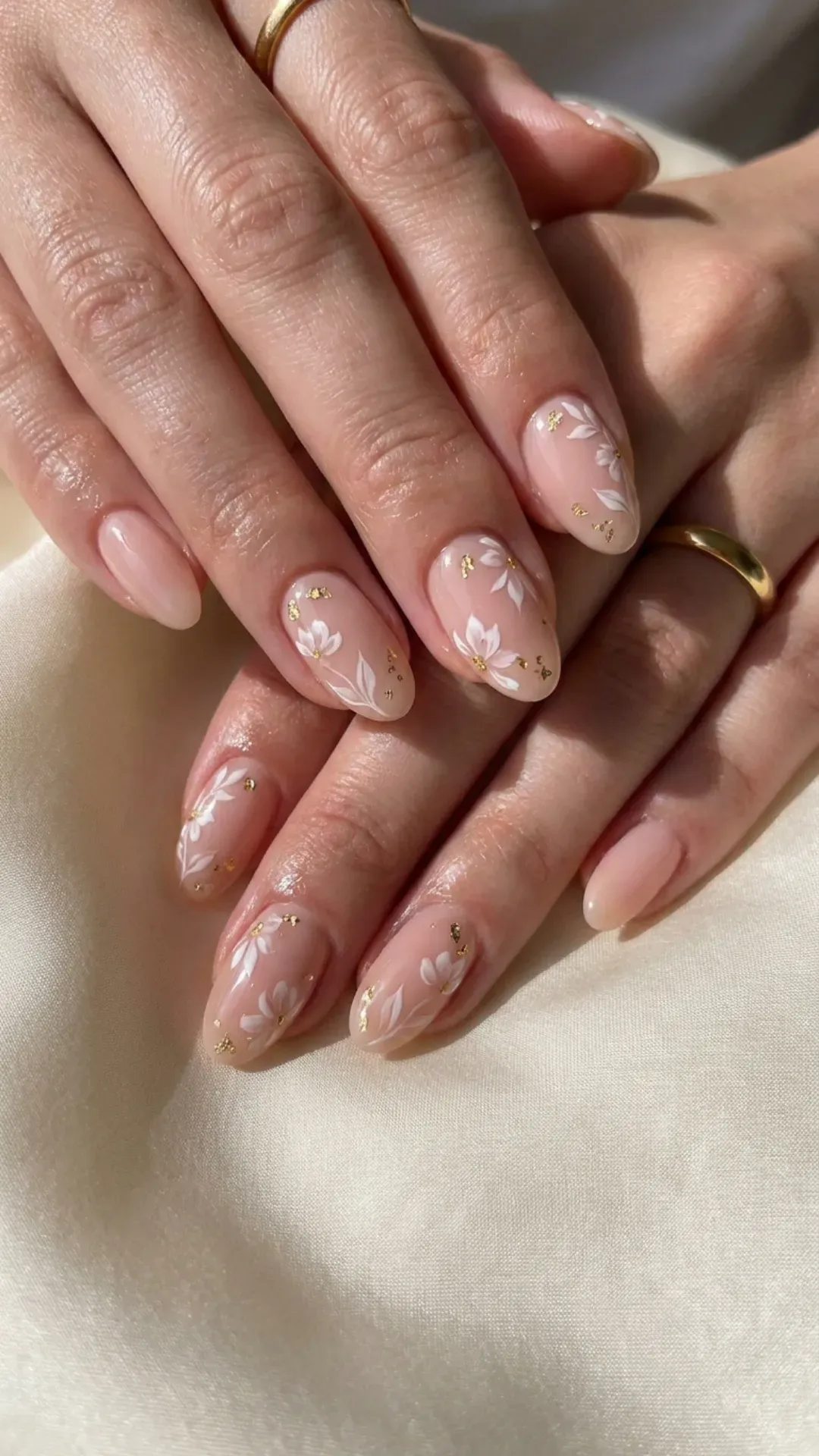 Natural-looking nude pink nails with delicate white floral accents and tiny gold flake embellishments for an elegant touch.
