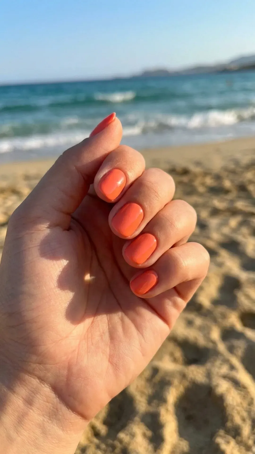 Short round nails in bright coral-orange high-gloss finish, ultra-shiny in warm sunlight, beach background — summer 2026